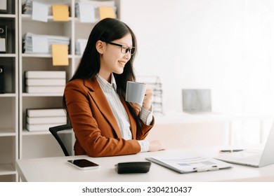 Confident beautiful Asian businesswoman typing laptop computer and digital tablet while holding coffee at modern office - Powered by Shutterstock
