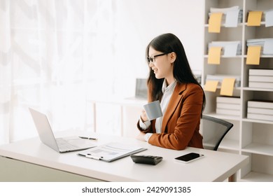 Confident beautiful Asian businesswoman typing laptop computer and digital tablet while holding coffee at modern office - Powered by Shutterstock