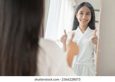 A confident and beautiful Asian woman in a bathrobe is smiling and giving herself a thumbs up in front of the mirror. Self-esteem, self-confidence, positive mindset, and self-motivation - Powered by Shutterstock