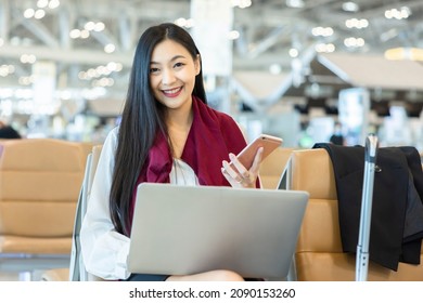 Confident Beautiful Young Chinese Woman Sitting On Bench At Airport Working Or Study On Laptop And Smartphone. Concept Travel, Remote Work And Education  