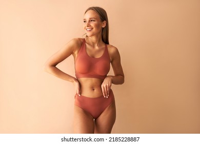 Confident beautiful young woman posing on beige background in comfortable underwear smiling and looking away. 