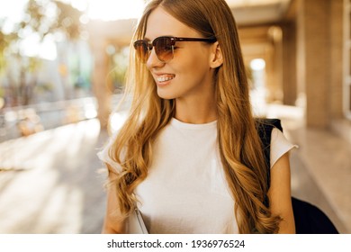 Confident beautiful young woman in sunglasses, in a white t-shirt is walking with a laptop in the city on the background of buildings