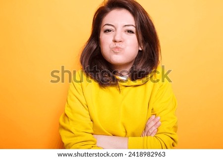 Confident beauty model with brunette hair makes a kissy face on camera while posing carefree in the studio. Portrait of young female adult with arms crossed making a goofy face.