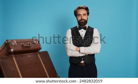 Confident bellboy posing on camera, wearing formal attire while he works at luxurious hotel. Staff member standing next to pile of suitcases and trolley bags, provide excellent concierge service.