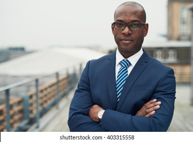 Confident black business man in a stylish suit standing with folded arms on a rooftop of n office block looking at the camera with a serious expression