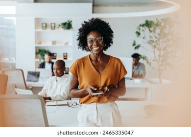 Confident black woman with afro hair stands in a modern office, happily engaged with her smartphone. She exudes professionalism and creativity, embodying the success of a young business person. - Powered by Shutterstock