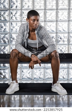 Confident Black woman in buzz cut hairstyle with earbuds, looking at camera, sitting on a bench with her elbows resting on her knees, against glass block wall. Full-length vertical