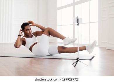 Confident black woman doing abs exercises on floor yoga mat, female fitness blogger shooting online tutorials, showing cross body twist knee to elbow crunches with arms behind head, watching video - Powered by Shutterstock