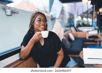 Confident Black woman with glasses and braids holding coffee cup, smiling warmly in cozy cafe corner near laptop and notes, combining remote work with peaceful personal moment - Powered by Shutterstock - Get 15% off with code: PIKWIZARD15
