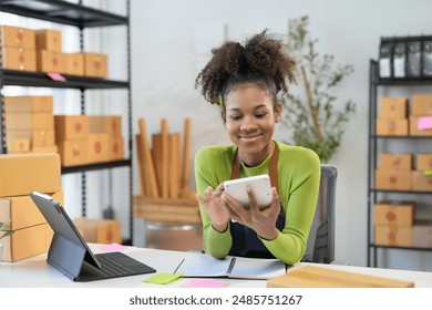 Confident black woman manages her small business from a warehouse office, calculating costs on a tablet with a smile - Powered by Shutterstock