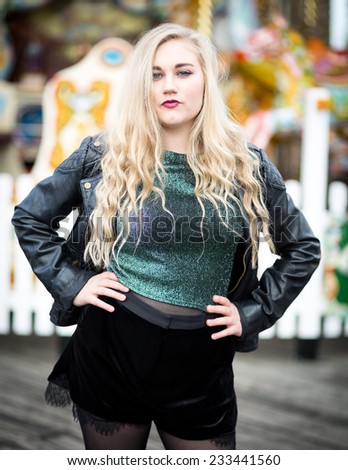 Confident blond teenager with long hair wearing a leather jacket, green glittery t-shirt, black shorts, nylons and high heeled boots standing in front of a fairground ride with a white fence around it