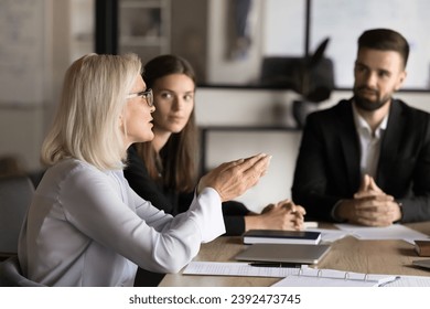 Confident blonde senior business professional woman talking to younger colleagues on brainstorming meeting, offering ideas for work on project, discussing strategy, management - Powered by Shutterstock