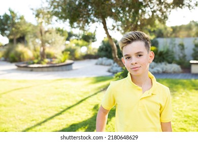 Confident Boy 8 Years Old With A Stylish Haircut And An Earring, Wearing A Yellow Polo, Standing In The Park. Green Grass On The Background. Sunny Summer Day. Concept Children, Kid, Childhood, Haircut