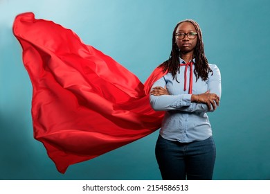 Confident And Brave Young Superhero Woman Standing With Arms Crossed While Wearing Red Mighty Cape On Blue Background. Portrait Of Justice Defender Posing Tough And Strong While Looking At Camera.