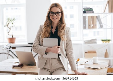Confident Business Expert. Attractive Young Smiling Woman In Smart Casual Wear Holding Digital Tablet And Looking At Camera While Leaning On Desk In Creative Office