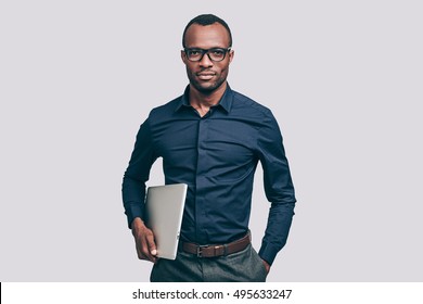 Confident business expert. Handsome young African man carrying laptop and looking at camera while standing against grey background