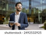 Confident business man in glasses and suit using tablet outdoors with modern office building in background. Concept of professional success, technology, and business lifestyle.