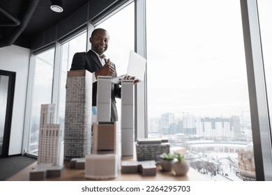 Confident business man real estate agents in black stylish formal suit standing with laptop near 3d model skyscrapers of city architecture design at office with panoramic city view, showing thumb up