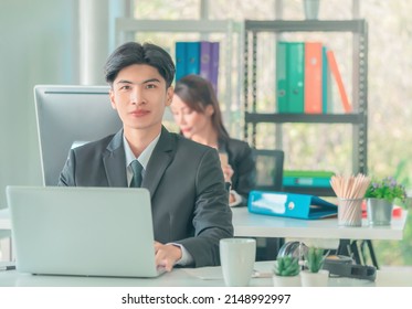 Confident Business Man Sitting Working With Laptop Computer In Office, Front View With Team In Background.