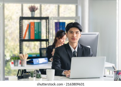 Confident Business Man Sitting Working With Laptop Computer In Office, Front View With Team In Background.