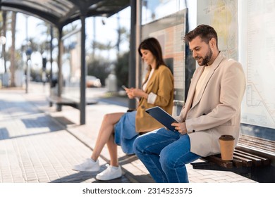 Confident business man with takeaway coffee sitting at bus station with a female, waiting for bus, using digital tablet device to check bus schedules, connections and maps, buying online bus ticket. - Powered by Shutterstock