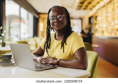 Confident business owner looking at the camera while working in a cafe. Mature businesswoman with dreadlocks using a laptop while working remotely. - Powered by Shutterstock