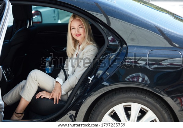 confident business woman inside of car, elegant\
blonde lady feel safety and comfort, in luxurious taxi, she looks\
at camera and smile