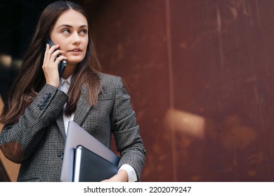 Confident Business Woman Walking, Holding Textbook, Laptop, Talking On Mobile Phone On City Street In Front Of Modern Office Building. Copy Space. Modern