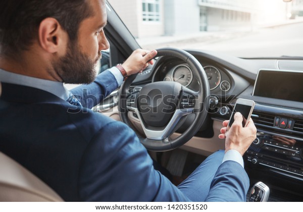 Confident businessman. Back view of confident and\
young bearded man in a stylish suit touching his smart phone while\
sitting in the car