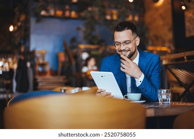 Confident businessman in blue suit using a digital tablet while sitting in a stylish cafe, enjoying coffee and working. Displays modern lifestyle and technology in a relaxed environment. - Powered by Shutterstock