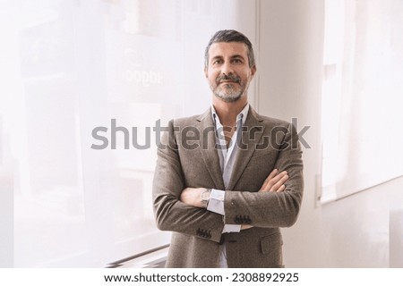 Confident Businessman in Bright Office Setting - Portrait of mature businessman with crossed arms, standing in white office illuminated by large window, in his forties, looking at camera