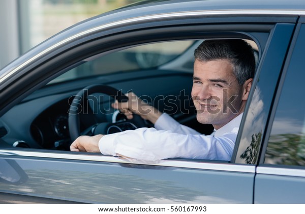 Confident businessman driving an expensive car, he\
is smiling at camera