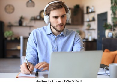 Confident businessman in earphones is writing notes or financial report while sitting at desk with laptop at home