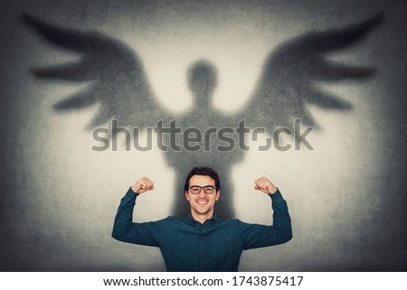 Confident businessman flexing muscles imagine superpower. Guy shows his strength, casting a superhero shadow with angel wings on a wall. Personal development, inner power and motivation concept.