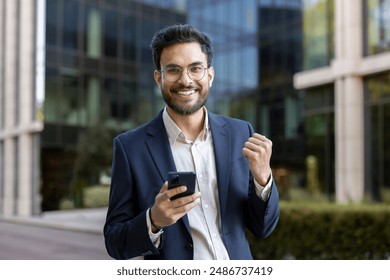 Confident businessman in formal attire standing outside modern office building, smiling at camera while using smartphone. Expressing joy and accomplishment, symbolizes success. - Powered by Shutterstock