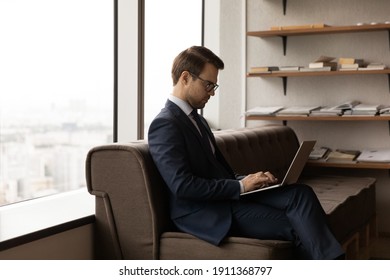 Confident Businessman In Formal Suit And Glasses Work On Laptop Hold Device On Knees While Expecting For Business Meeting. Serious Young Man Visitor Sit On Sofa In Office Waiting Room Using Computer