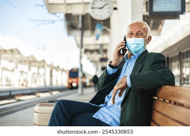 Confident businessman in protective face mask, talking on mobile phone while sitting outside the train station with a suitcase. - Powered by Shutterstock