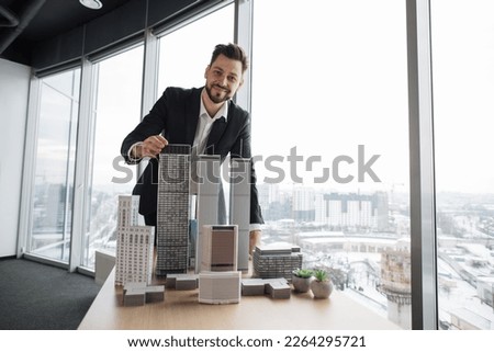 Confident businessman real estate agents in black stylish formal suit standing while touching 3d model skyscraper of city architecture design at office with panoramic city view. [[stock_photo]] © 