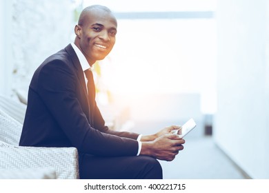 Confident businessman. Side view of cheerful young African businessman working on digital tablet and smiling at camera