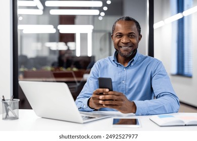 Confident businessman smiling while using smartphone at modern office desk. Professional workspace with laptop, tablet, and office supplies. Concept of business, technology, and corporate lifestyle. - Powered by Shutterstock