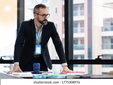 Confident Businessman Standing At The Table And Two His Hand Touch On The Messy Table In The Modern Office At A High Level With Blurred Background. He Is Not Looking At The Camera. 