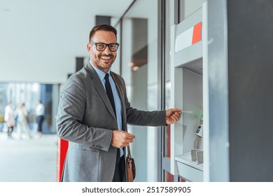 Confident businessman using an ATM outside, smiling at the camera. The setting features modern office buildings with people walking in the background, emphasizing convenience and technology. - Powered by Shutterstock