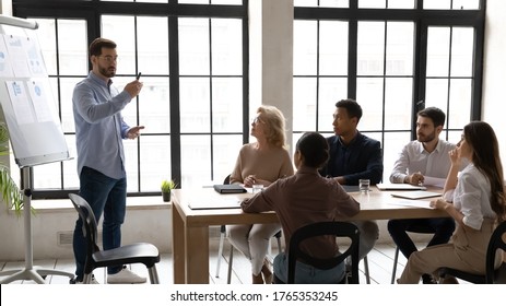 Confident businessman wearing glasses leading corporate meeting with diverse staff, making flip chart presentation, business coach mentor training employees, explaining strategy, presenting stats