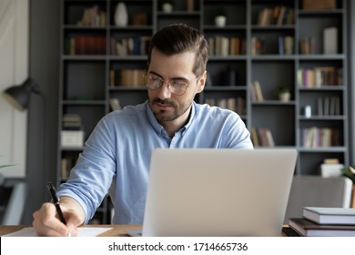 Confident businessman wearing glasses writing notes or financial report, sitting at desk with laptop, focused serious man working with paper documents, student studying online, research work - Powered by Shutterstock