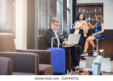 Confident Businessman Working With Laptop At The Airport Lounge. Man Resting During Waiting His Flight With Suitcase And Blurred Background Of Passengers Enjoying Drink At Counter Serving Drinks.  