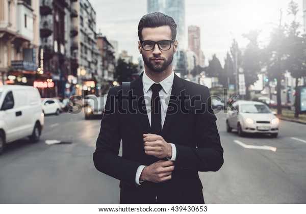 Confident businessman. Confident young man in\
full suit adjusting his sleeve and looking away while standing\
outdoors with cityscape in the\
background