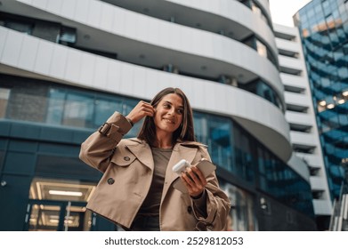 Confident businesswoman adjusts earbuds, walks city streets with smartphone, listening to music, embodying tech savvy style - Powered by Shutterstock