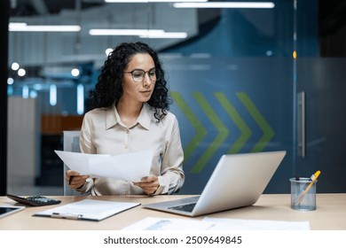 Confident businesswoman analyzing documents while using laptop in modern office. Casual attire suggests comfortable work setting. Emphasis on focus and productivity. - Powered by Shutterstock