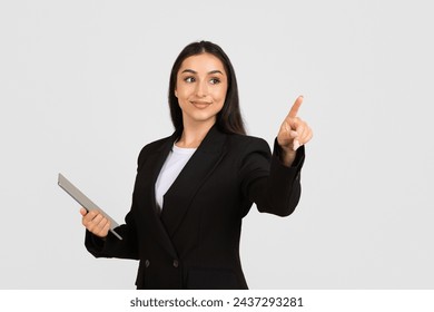 Confident businesswoman in black suit with digital tablet making pointing gesture as if interacting with an invisible touchscreen over light background - Powered by Shutterstock