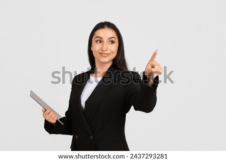 Confident businesswoman in black suit with digital tablet making pointing gesture as if interacting with an invisible touchscreen over light background